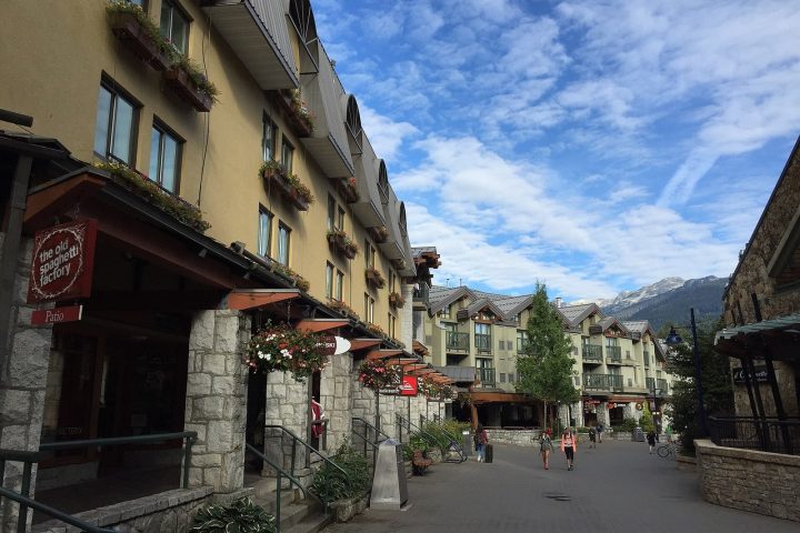 The pedestrian Village Stroll in Whistler Village, lined with stone-fronted shops and flower boxes, with snow on the peaks beyond