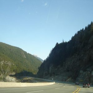 Highway 99 curving through a forested canyon near Squamish, with a power line tower and rock cut beside the road