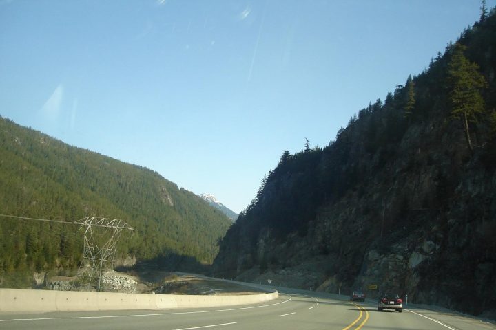 Highway 99 curving through a forested canyon near Squamish, with a power line tower and rock cut beside the road